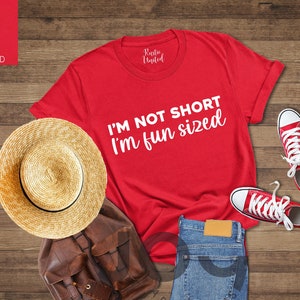 May include: Red t-shirt with the text "I'm not short I'm fun sized" in white lettering. The shirt is laid out on a wooden surface with a straw hat, a brown leather backpack, and red sneakers.