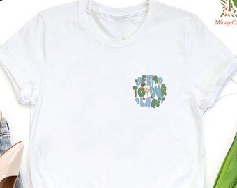 May include: White t-shirt with a graphic design that says "Be Kind To Our Planet" in a colorful, whimsical font. The graphic is surrounded by small flowers and leaves.