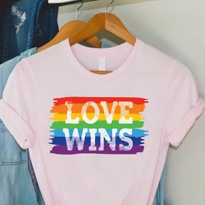 May include: A light pink t-shirt with the words "LOVE WINS" in white letters over a rainbow-colored horizontal stripe design. The shirt is hanging on a wooden hanger.