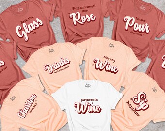 May include: A set of nine t-shirts with various wine-themed slogans printed on them. The shirts are in two colours: peach and maroon. The slogans include "Corks are for quitters", "Back that glass up", "Stop and smell the Rose", "Drinks well with others", "Rise and Wine", "I make Pour decisions", "Bride on cloud wine", "Partners in Wine", and "Sip happens".