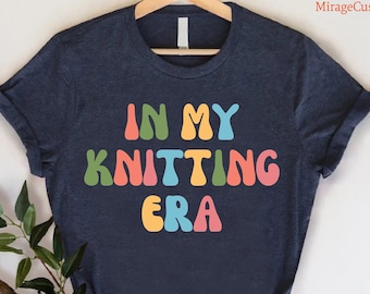May include: A navy blue t-shirt with the text "In My Knitting Era" in colorful, retro-style lettering.