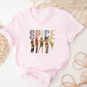 May include: A light pink t-shirt featuring the word "SPICE" in a colorful, pixelated font. Below the text are illustrations of five women in various outfits. The t-shirt is made of a soft material.