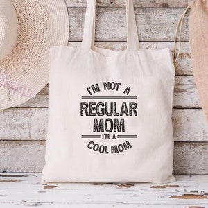 May include: Cream-colored canvas tote bag with the text "I'M NOT A REGULAR MOM I'M A COOL MOM" in black. The bag has two handles and is displayed against a rustic, white-washed wooden background. A straw hat and a vase of flowers are also visible.