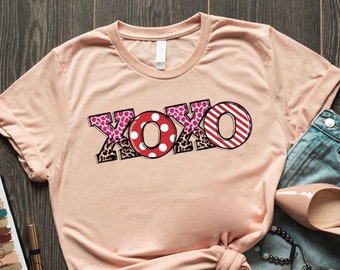 May include: A peach-colored t-shirt with a graphic print of the word "XOXO" in pink, red, and black patterned letters. The shirt is folded and laid on a wooden surface with a pair of light brown high heels, blue jeans, and makeup products.