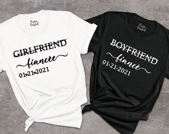 May include: Two white t-shirts with black text. One shirt says "Girlfriend fiance 01.21.2021" and the other says "Boyfriend fiance 01.21.2021".