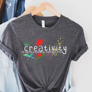 May include: A dark gray t-shirt with a white graphic that says "creativity is intelligence having fun." The graphic is surrounded by colorful paint splatters.