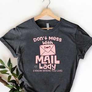 May include: A dark gray t-shirt with a pink graphic that says "Don't mess with Mail Lady (I know where you live)". The graphic features a cartoon envelope with a smiling face.