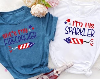 May include: Two t-shirts with a Fourth of July theme. The blue shirt says "She's my firecracker" with a red, white, and blue firework graphic. The white shirt says "I'm his sparkler" with a red, white, and blue firework graphic.