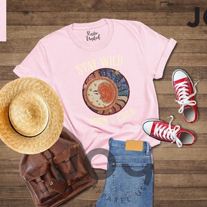 May include: Pink t-shirt featuring a graphic of a crescent moon and stars with the text "Stay Wild Moon Child". The shirt is laid out on a wooden surface with a straw hat, a brown leather backpack, and red trainers.