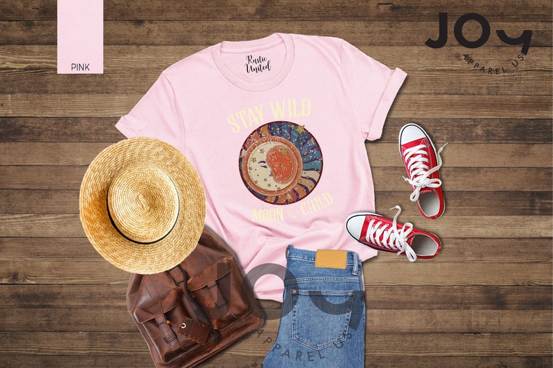 May include: Pink t-shirt featuring a graphic of a crescent moon and stars with the text "Stay Wild Moon Child". The shirt is laid out on a wooden surface with a straw hat, a brown leather backpack, and red trainers.