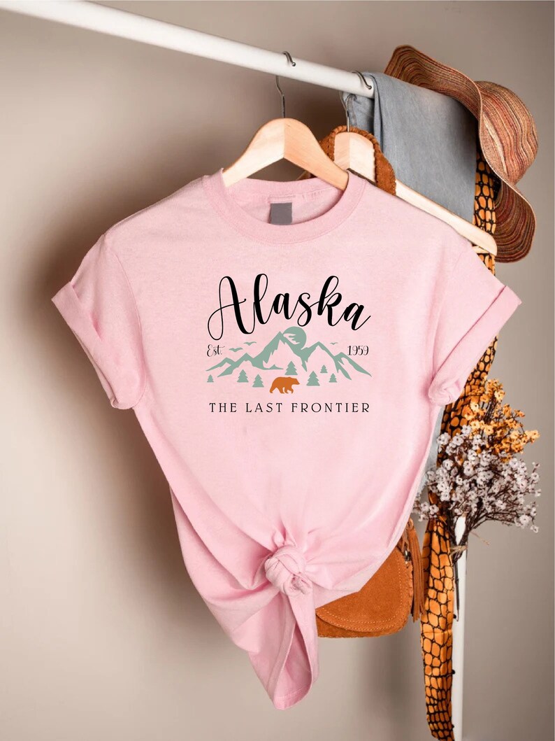 May include: Pink t-shirt with a graphic design of mountains and a bear. The text "Alaska" is written in a cursive font above the mountains. The text "Est. 1959" is written below the mountains. The text "The Last Frontier" is written below the mountains.