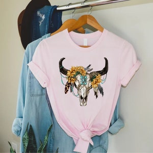 K&ouml;nnte beinhalten: Ein pinkfarbenes T-Shirt mit einem wei&szlig;en Totenkopf-Design. Der Totenkopf ist mit Sonnenblumen, t&uuml;rkisfarbenen Perlen und Federn verziert.