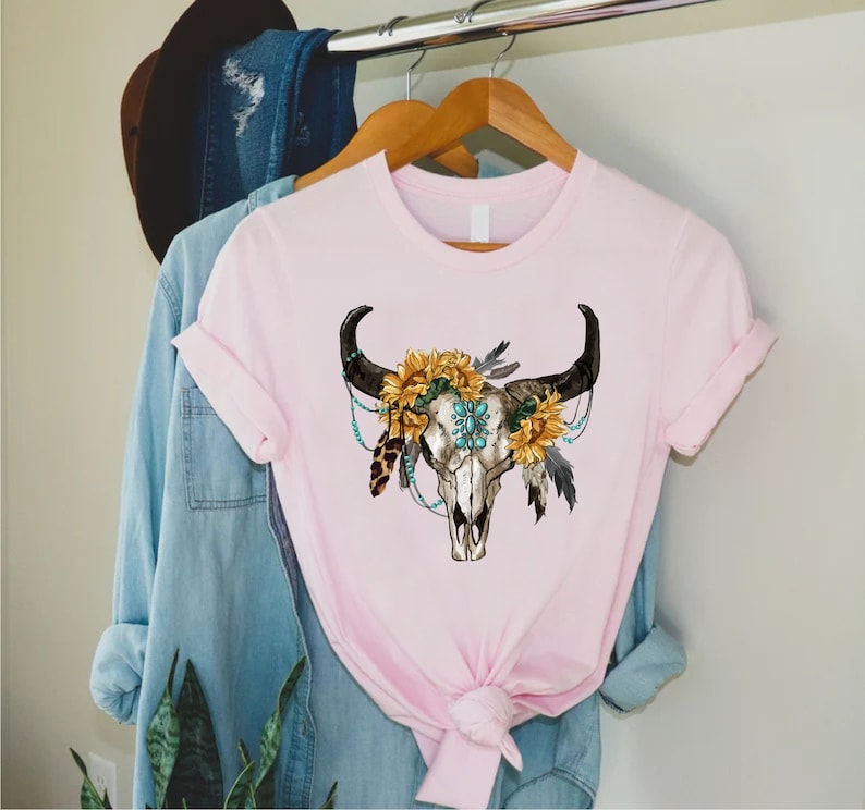 K&ouml;nnte beinhalten: Ein pinkfarbenes T-Shirt mit einem wei&szlig;en Totenkopf-Design. Der Totenkopf ist mit Sonnenblumen, t&uuml;rkisfarbenen Perlen und Federn verziert.