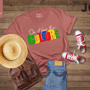 May include: A heather mauve t-shirt with the text "Do it for the CULTURE" in colorful block letters. The shirt is laid out on a wooden surface with a straw hat, a brown leather backpack, a pair of red sneakers, and blue jeans.