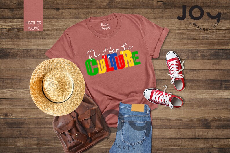 May include: A heather mauve t-shirt with the text "Do it for the CULTURE" in colorful block letters. The shirt is laid out on a wooden surface with a straw hat, a brown leather backpack, a pair of red sneakers, and blue jeans.
