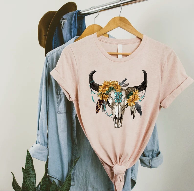 K&ouml;nnte beinhalten: Ein pinkfarbenes T-Shirt mit einem Aufdruck eines wei&szlig;en Bullensch&auml;dels, der mit Sonnenblumen, t&uuml;rkisfarbenen Perlen und Federn verziert ist.
