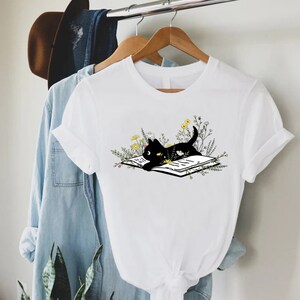 May include: White t-shirt with a black cat reading a book surrounded by flowers.