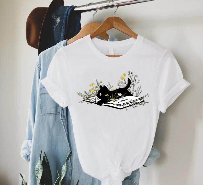 May include: White t-shirt with a black cat reading a book surrounded by flowers.