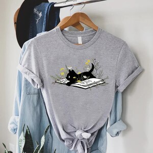 May include: A gray t-shirt with a black cat reading a book surrounded by flowers. The cat is looking at the book.