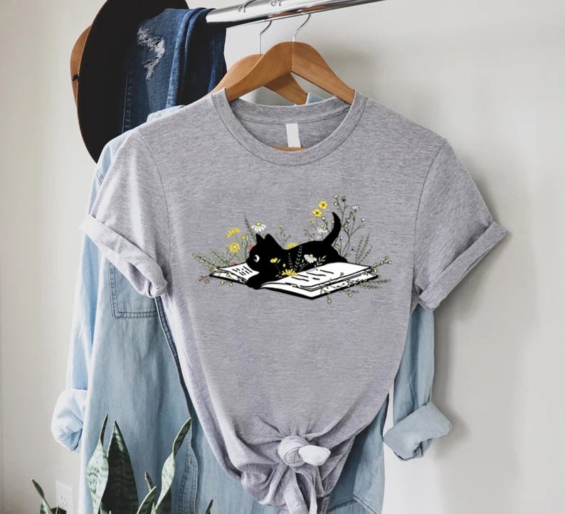 May include: A gray t-shirt with a black cat reading a book surrounded by flowers. The cat is looking at the book.