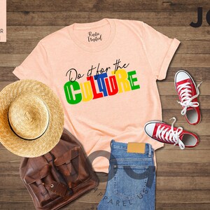 May include: A peach colored t-shirt with the text "Do it for the CULTURE" in colorful block letters. The shirt is laid out on a wooden surface with a straw hat, a brown leather backpack, a pair of blue jeans, and red sneakers.