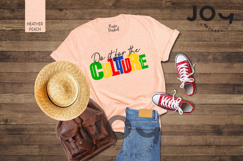 May include: A peach colored t-shirt with the text "Do it for the CULTURE" in colorful block letters. The shirt is laid out on a wooden surface with a straw hat, a brown leather backpack, a pair of blue jeans, and red sneakers.