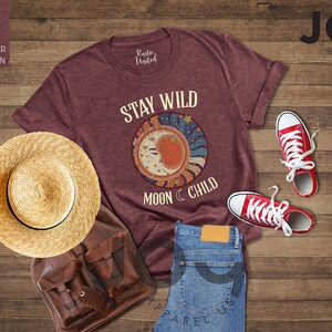 May include: A heather maroon t-shirt featuring the text "Stay Wild Moon Child" and a graphic of a crescent moon with stars. The shirt is laid out on a wooden surface with a straw hat, a leather bag, and red trainers.