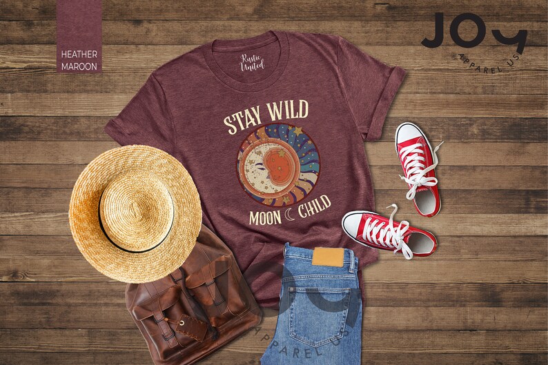 May include: A heather maroon t-shirt featuring the text "Stay Wild Moon Child" and a graphic of a crescent moon with stars. The shirt is laid out on a wooden surface with a straw hat, a leather bag, and red trainers.