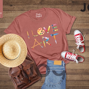 Puede incluir: Una camiseta de color malva jaspeado con las palabras "ME ENCANTA EL ARTE" escritas con pinceles, l&aacute;pices y una paleta de pintura. La camiseta est&aacute; colocada sobre una superficie de madera con un sombrero de paja, una mochila de cuero marr&oacute;n, unos vaqueros azules y unas zapatillas rojas.