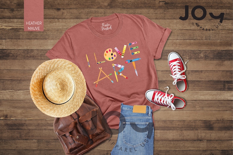 Puede incluir: Una camiseta de color malva jaspeado con las palabras "ME ENCANTA EL ARTE" escritas con pinceles, l&aacute;pices y una paleta de pintura. La camiseta est&aacute; colocada sobre una superficie de madera con un sombrero de paja, una mochila de cuero marr&oacute;n, unos vaqueros azules y unas zapatillas rojas.