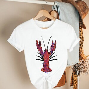 May include: White t-shirt with a red and purple crayfish illustration. The crayfish is outlined in black and has a watercolor effect.