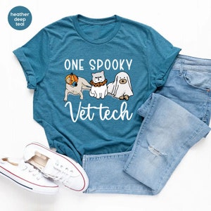 May include: A heather deep teal t-shirt featuring a graphic of a dog wearing a pumpkin, a cat with a collar, and a ghost. The text reads ONE SPOOKY Vet tech. The shirt is paired with light blue distressed denim jeans and white canvas sneakers.
