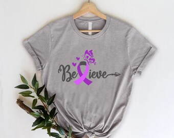 May include: A gray t-shirt with the word "Believe" in a script font, a purple awareness ribbon, and a butterfly graphic. The shirt is displayed on a wooden hanger.