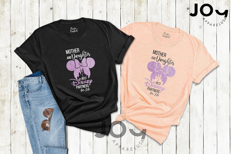 Puede incluir: Dos camisetas, una negra y otra melocot&oacute;n, con el texto "Mother and Daughter Best Disney Partners for Life". El dise&ntilde;o presenta una silueta de Minnie Mouse con un castillo. Tambi&eacute;n se ven unos vaqueros azules y gafas de sol.