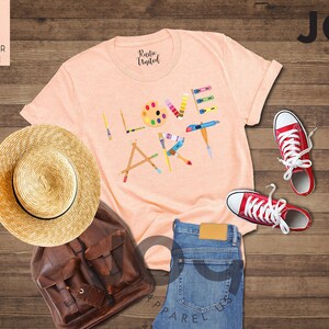 Puede incluir: Una camiseta de color melocot&oacute;n con el texto "I Love Art" en letras de colores formadas por materiales de arte. La camiseta est&aacute; colocada sobre una superficie de madera con un sombrero de paja, una mochila de cuero marr&oacute;n y zapatillas rojas.