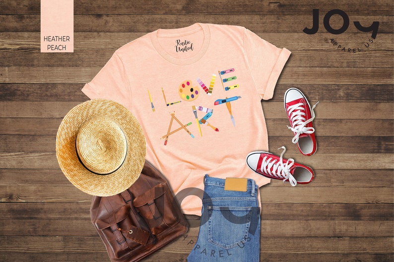 Puede incluir: Una camiseta de color melocot&oacute;n con el texto "I Love Art" en letras de colores formadas por materiales de arte. La camiseta est&aacute; colocada sobre una superficie de madera con un sombrero de paja, una mochila de cuero marr&oacute;n y zapatillas rojas.