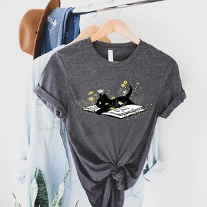 May include: A dark gray t-shirt with a black cat reading a book surrounded by yellow flowers. The cat is lying on the book with its head resting on the page.