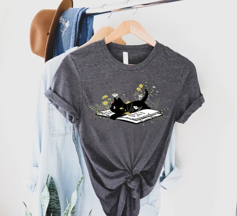 May include: A dark gray t-shirt with a black cat reading a book surrounded by yellow flowers. The cat is lying on the book with its head resting on the page.
