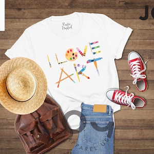 Puede incluir: Camiseta blanca con el texto "I LOVE ART" en letras de colores hechas de pinceles, l&aacute;pices y una paleta de pintura. La camiseta est&aacute; colocada sobre una superficie de madera con un sombrero de paja, una mochila de cuero marr&oacute;n, unos vaqueros azules y unas zapatillas rojas.
