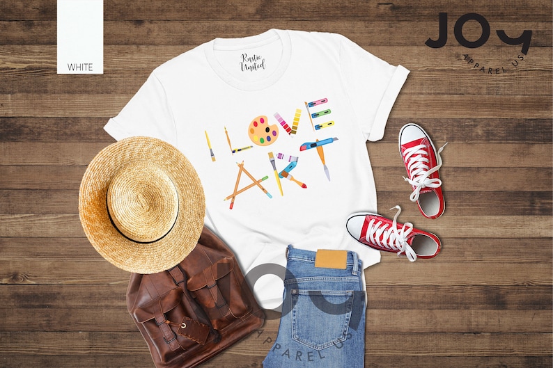 Puede incluir: Camiseta blanca con el texto "I LOVE ART" en letras de colores hechas de pinceles, l&aacute;pices y una paleta de pintura. La camiseta est&aacute; colocada sobre una superficie de madera con un sombrero de paja, una mochila de cuero marr&oacute;n, unos vaqueros azules y unas zapatillas rojas.