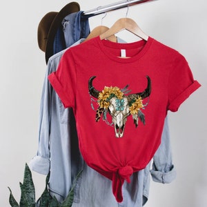 K&ouml;nnte beinhalten: Rotes T-Shirt mit einem Aufdruck eines wei&szlig;en Bullensch&auml;dels mit einer t&uuml;rkisfarbenen und goldenen Blumenkrone. Der Sch&auml;del ist von Federn und Perlen umgeben.