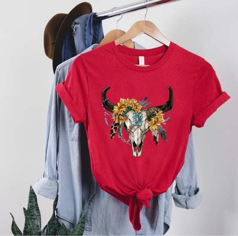 K&ouml;nnte beinhalten: Rotes T-Shirt mit einem Aufdruck eines wei&szlig;en Bullensch&auml;dels mit einer t&uuml;rkisfarbenen und goldenen Blumenkrone. Der Sch&auml;del ist von Federn und Perlen umgeben.