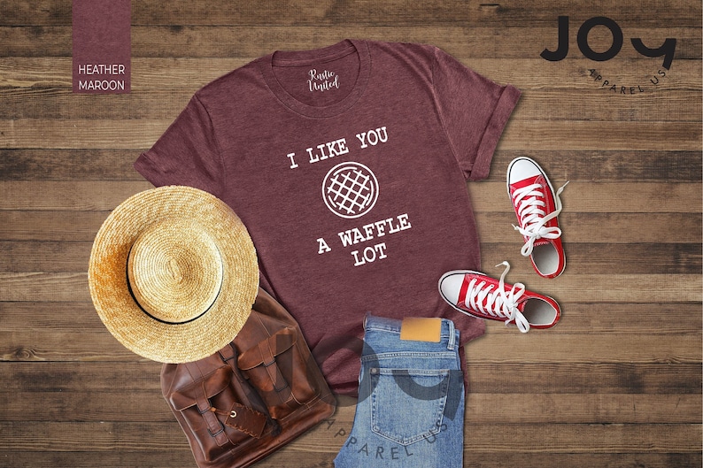 Puede incluir: Una camiseta de color marr&oacute;n jaspeado con el texto "I like you a waffle lot" y un gr&aacute;fico de waffle. La camiseta est&aacute; colocada sobre una superficie de madera con un sombrero de paja, una bolsa de cuero marr&oacute;n y zapatillas rojas.