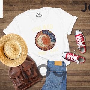 May include: White t-shirt featuring a graphic of a moon and stars with the text "Stay Wild Moon Child". The shirt is laid out on a wooden surface with a straw hat, a brown leather backpack, blue jeans, and red trainers.