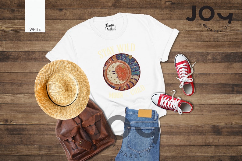 May include: White t-shirt featuring a graphic of a moon and stars with the text "Stay Wild Moon Child". The shirt is laid out on a wooden surface with a straw hat, a brown leather backpack, blue jeans, and red trainers.