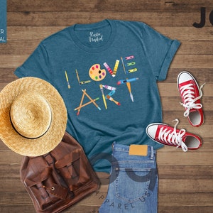 Puede incluir: Una camiseta azul turquesa con las palabras "I LOVE ART" en letras de colores. Las letras est&aacute;n formadas por diversos materiales de arte, como pinceles, l&aacute;pices y una paleta de pintura. La camiseta est&aacute; colocada sobre una superficie de madera con un sombrero de paja, una mochila de cuero marr&oacute;n y un par de zapatillas rojas.