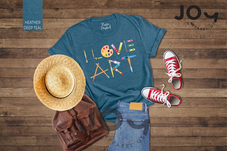 Puede incluir: Una camiseta azul turquesa con las palabras "I LOVE ART" en letras de colores. Las letras est&aacute;n formadas por diversos materiales de arte, como pinceles, l&aacute;pices y una paleta de pintura. La camiseta est&aacute; colocada sobre una superficie de madera con un sombrero de paja, una mochila de cuero marr&oacute;n y un par de zapatillas rojas.