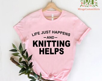 May include: Pink t-shirt with the text "Life just happens -and- Knitting helps" in black.