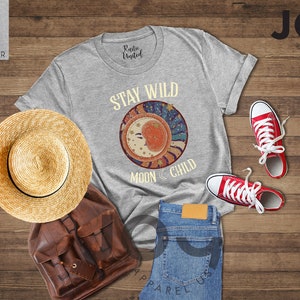 May include: A light grey heather t-shirt with the text "Stay Wild Moon Child" and a graphic of a crescent moon with stars and a sunburst design.