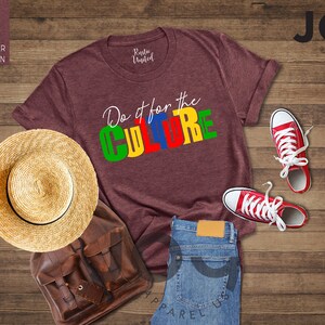 May include: Heather maroon t-shirt with the text "Do it for the CULTURE" in colorful block letters. The shirt is laid out on a wooden surface with a straw hat, a brown leather backpack, a pair of red sneakers, and blue jeans.